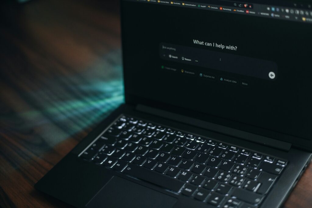 A close-up of a laptop with a dark interface displaying the prompt “What can I help with?” on screen.
